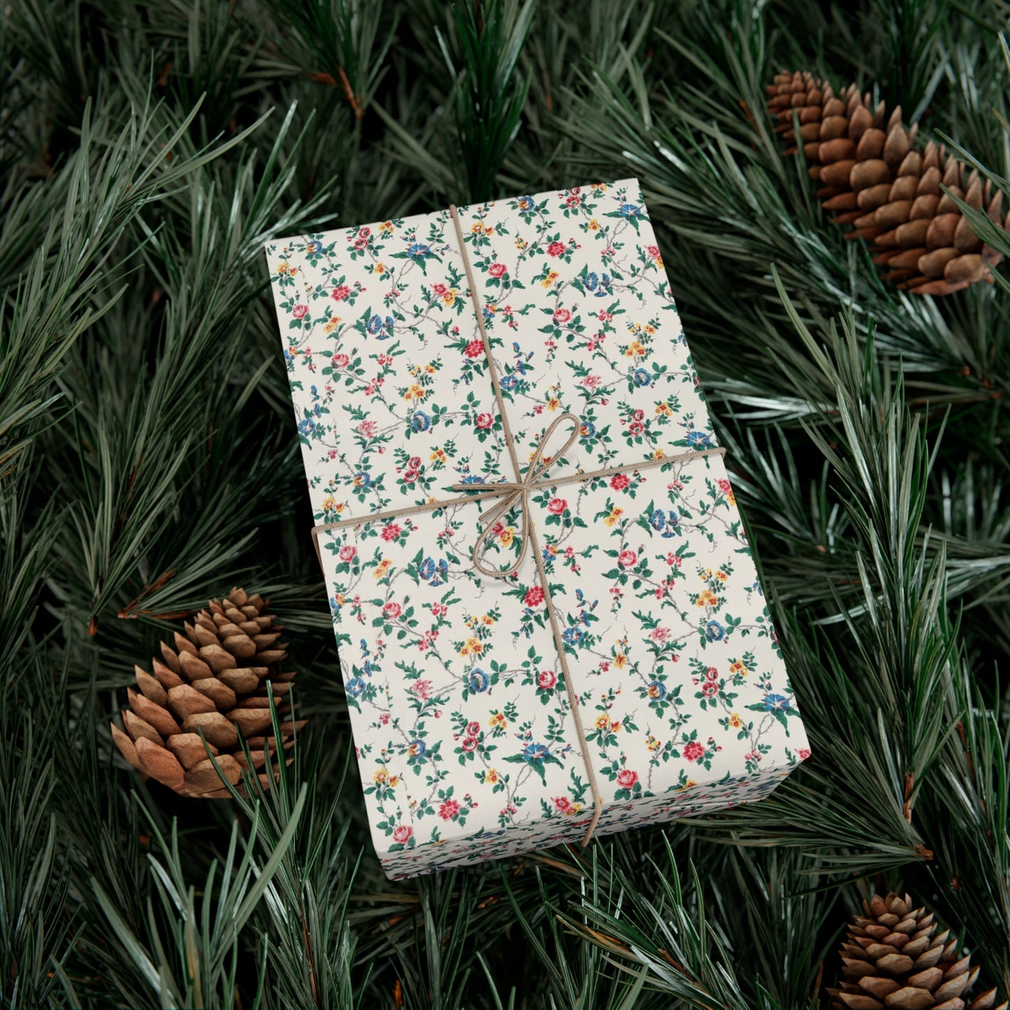 Gift box wrapped in floral paper with brown string on a bed of pine needles and pine cones.